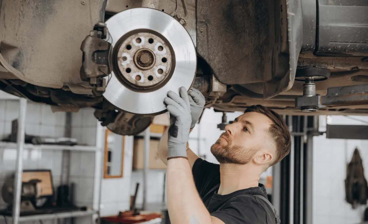 A mechanic wearing gloves inspects a European cars brake system in a Guelph garage. The vehicle is elevated on a hydraulic lift, revealing the shiny brake disc. Shelves filled with tools and equipment are visible in the background. Euro Mechanic | Brock Road Garage Guelph | Top Reviewed Auto Repair Shop in Guelph | Visit brockroadgarage.ca