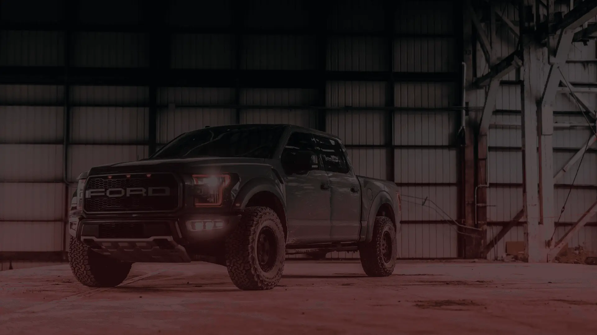 A large black Ford pickup truck with off-road tires is parked inside a dimly lit industrial warehouse, Guelphs finest mechanic shop, with metal beams and a corrugated wall in the background. The headlights are on, casting a glow on the ground. EuroMechanic | Brock Road Garage Guelph | Top Reviewed Auto Repair Shop in Guelph | Visit brockroadgarage.ca