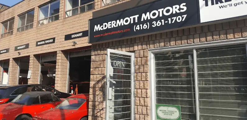 Exterior of McDermott Motors, an auto repair shop resembling the charm of Brock Road Garage. Beige brick walls house a sign with the name and phone number. The entrance door reads OPEN, while a red convertible is parked nearby. Garage bays are visible on the left side, inviting vehicles in from all locations. Euro Mechanic | Brock Road Garage Guelph | Top Reviewed Auto Repair Shop in Guelph | Visit brockroadgarage.ca