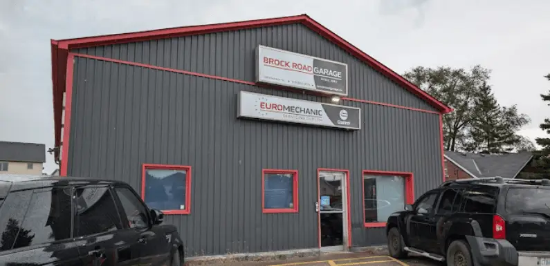 The small gray garage building, marked by its red trim and Brock Road Garage and Euromechanic signs, is nestled in a quaint location. Two cars are parked out front, with lush trees serving as a backdrop. Euro Mechanic | Brock Road Garage Guelph | Top Reviewed Auto Repair Shop in Guelph | Visit brockroadgarage.ca