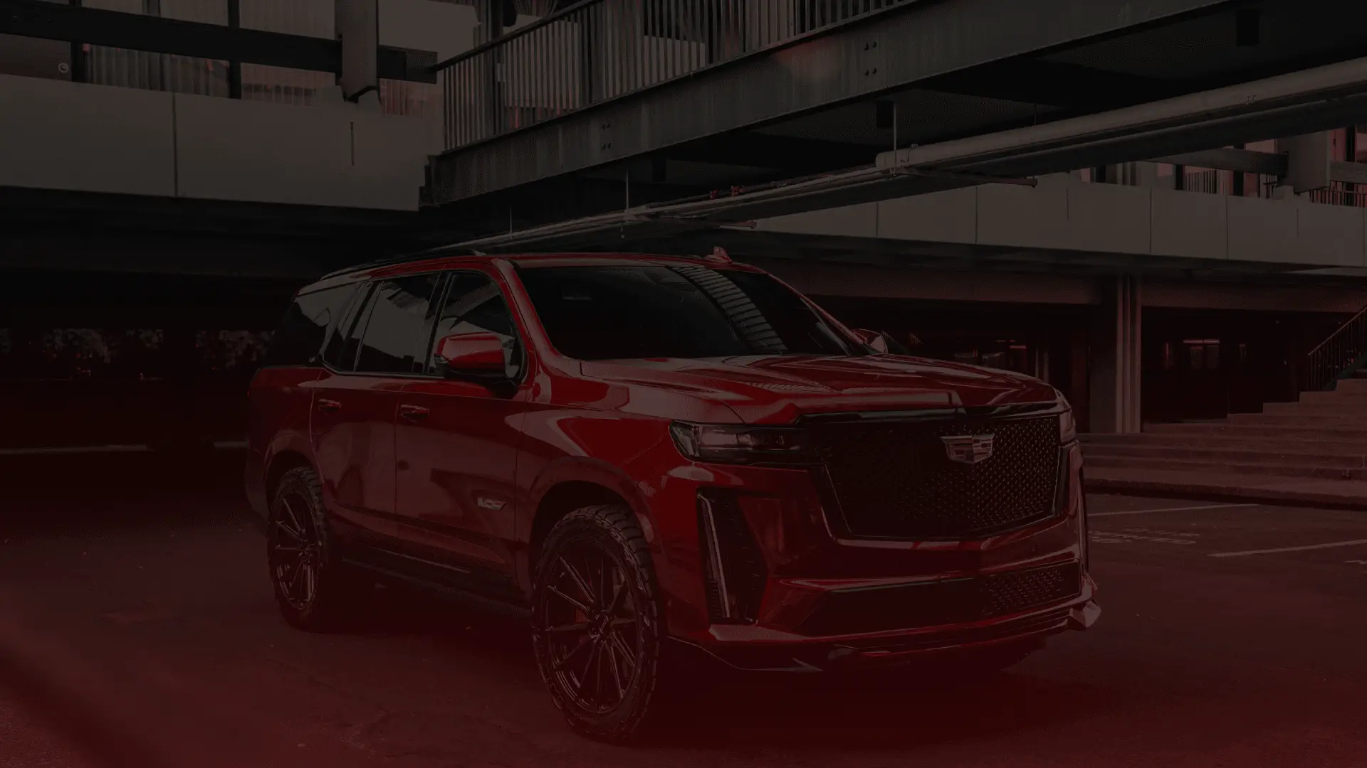 A sleek, red Cadillac SUV, perhaps just serviced by a Cadillac mechanic in Guelph, is parked in an urban setting under a modern multi-level structure. The cars glossy paint and intricate design details are highlighted, creating a striking contrast with the concrete surroundings. EuroMechanic | Brock Road Garage Guelph | Top Reviewed Auto Repair Shop in Guelph | Visit brockroadgarage.ca