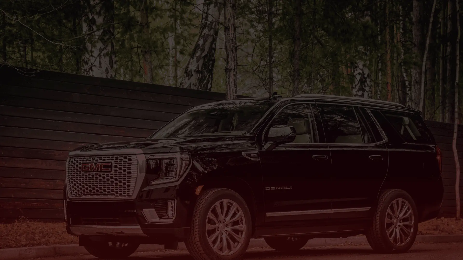 A black GMC Denali SUV is parked on the road in front of a dark wooden fence, surrounded by trees. Its sleek design and polished finish highlight luxury and ruggedness, reminding one of the expert care it might receive from a GM mechanic in Guelph. EuroMechanic | Brock Road Garage Guelph | Top Reviewed Auto Repair Shop in Guelph | Visit brockroadgarage.ca
