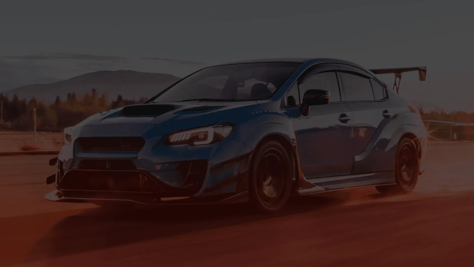 A blue sports car, reminiscent of the precision-tuned vehicles a Subaru mechanic in Guelph might perfect, speeds down a track at sunset. It raises dust, boasting a large rear spoiler and aggressive styling. Blurred mountains and trees frame the scene under a pinkish-orange sky. EuroMechanic | Brock Road Garage Guelph | Top Reviewed Auto Repair Shop in Guelph | Visit brockroadgarage.ca