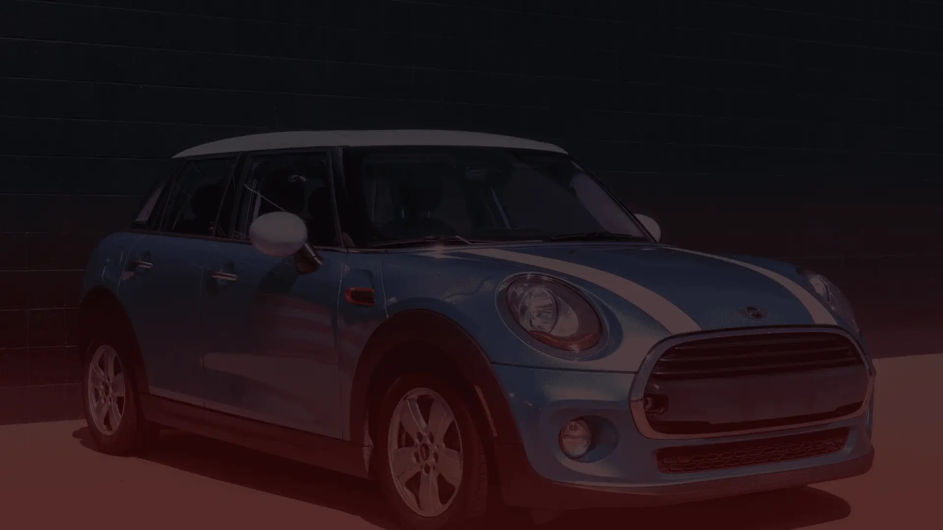 A blue Mini Cooper Guelph edition with a white roof and racing stripes is parked beside a dark brick wall. This four-door model, equipped with silver rims and rounded headlights, rests under a subtle dark red gradient overlay at the bottom. EuroMechanic | Brock Road Garage Guelph | Top Reviewed Auto Repair Shop in Guelph | Visit brockroadgarage.ca