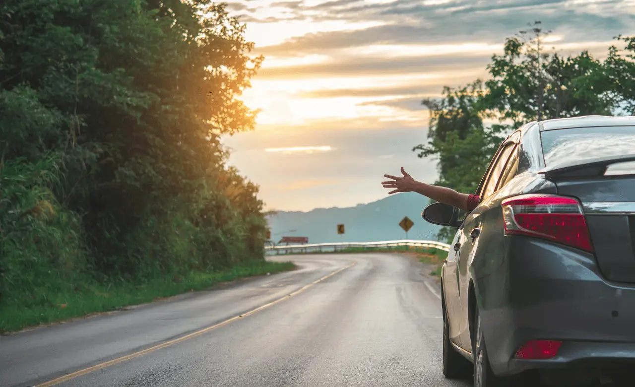 A car is driving along a scenic, winding road surrounded by lush greenery. The sun is setting in the background, casting a warm glow. A person inside the car is reaching out of the window with their arm extended. Euro Mechanic | Brock Road Garage Guelph | Top Reviewed Auto Repair Shop in Guelph | Visit brockroadgarage.ca